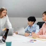 A group meeting with four colleagues around a table, two working on laptops. One person stands, speaking intensely. The mood is focused and professional.