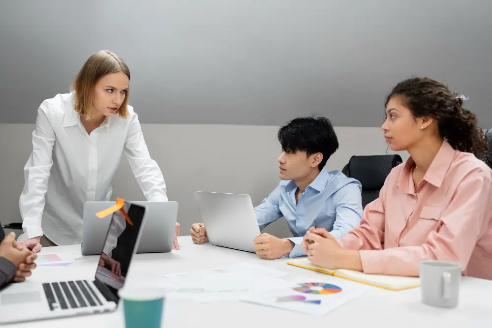 A group meeting with four colleagues around a table, two working on laptops. One person stands, speaking intensely. The mood is focused and professional.