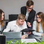 A group of four young professionals collaborating at a desk with laptops and documents. One stands pointing at a clipboard, fostering teamwork.