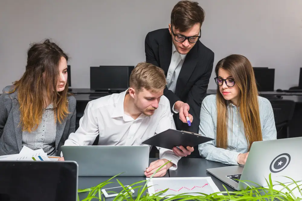 A group of four young professionals collaborating at a desk with laptops and documents. One stands pointing at a clipboard, fostering teamwork.