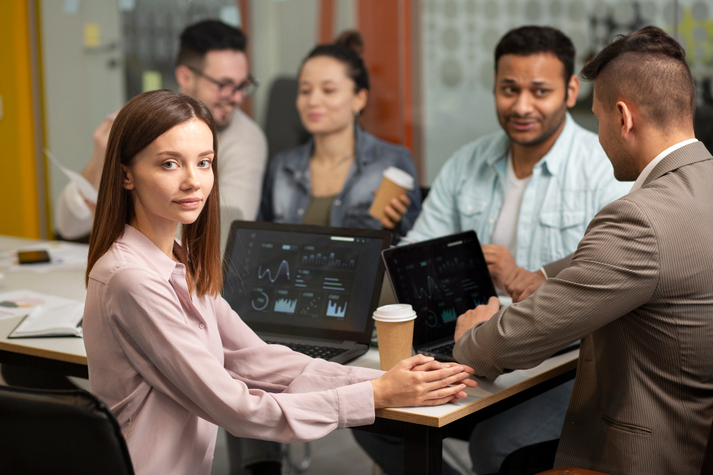A woman in a pink blouse sits at a table, facing the camera, amid a group discussion. Two laptops show graphs. The atmosphere is collaborative and focused.