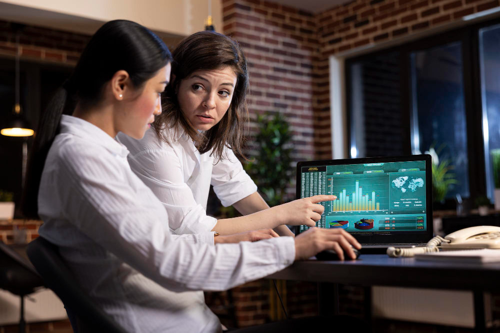 Two women in an office review data on a laptop screen displaying graphs and charts. They appear focused and engaged in discussion.